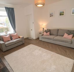 A living room with two gray sofas pink cushions a white rug wooden floor and blue curtains at Cosy Cottage in Amble
