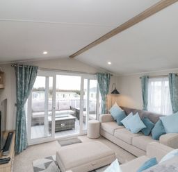 A living room with a beige sofa and ottoman with blue cushions and teal curtains by sliding glass doors opening to an outdoor seating area at Rowan Court 2 in Cayton