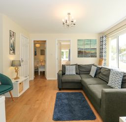 A living room with a grey sectional sofa and patterned cushions next to large windows at 1 The Dell in Mundesley