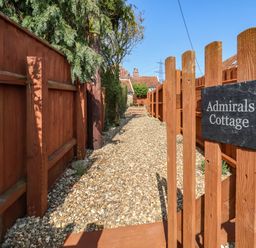 A gravel pathway with wooden fences on both sides and a wooden gate with a sign that reads Admirals Cottage at Admiral Cottage in Blaxhall near Snape Suffolk