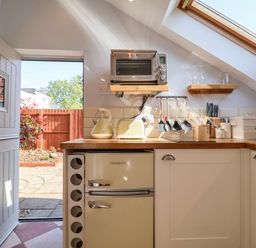 A kitchen with an open door showing the outside a small fridge kettle toaster utensils microwave and shelves at Admiral Cottage in Blaxhall near Snape Suffolk