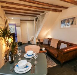 A living room with a dining table set for two a brown leather sofa plants and exposed wooden beams at Mulberry Tree in Appledore