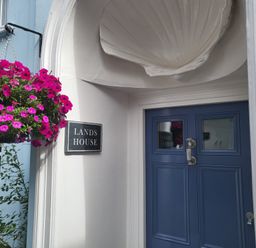 A blue door with silver handle under a white shell-shaped overhang next to a hanging basket of pink flowers at Lands House in Appledore