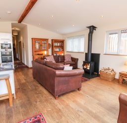 An open living room and kitchen area with brown leather sofas a wood burning stove a wooden floor and bookshelves at Gloccamaura in Bovey Tracey