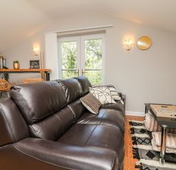 A living room with a brown leather sofa soft cushions a glass coffee table with a tray and throws a wooden floor near a dining area with wicker chairs and double doors at Hazelwood Lodge in High Bickington