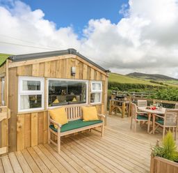 A wooden deck with outdoor seating and a dining table next to a wooden cabin surrounded by green hills at Caban Cwtch The Cosy Cabin in Llanaelhaearn