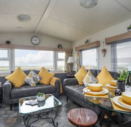 A living room with two grey sofas with yellow and patterned cushions a glass coffee table and a dining table set with yellow dishes at Caban Cwtch (The Cosy Cabin) in Llanaelhaearn