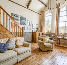 A sitting area with a beige sofa and armchair near a wooden staircase and a dining table with chairs by large windows at St Marks School House Nant-Glas near Rhayader