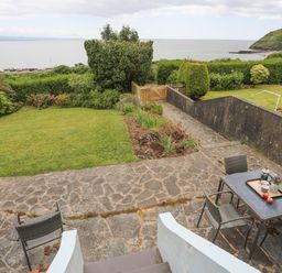 A garden with a stone patio and outdoor table with chairs overlooking the sea and a hill with a castle at Llys Owen in Criccieth