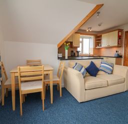A small dining area with wooden table and chairs next to a beige sofa with blue cushions in an open plan kitchen and living space at 5 Seymour Villas in Woolacombe