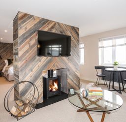 A living area with a wood-burning stove under a mounted TV a glass coffee table with books and a plant and a small dining table with two chairs near windows at The Hayloft in Llanfechell
