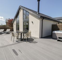 An outdoor patio area with a round table and four chairs two lounge chairs a hot tub and a building with large windows at Garden House in Llanfechell