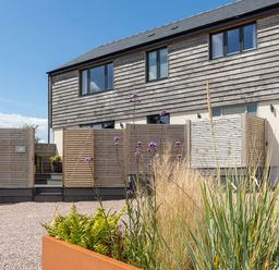 A modern two-story building with wooden cladding and a gravel courtyard with a planter of tall grasses and purple flowers at The Farmhouse in Llanfechell