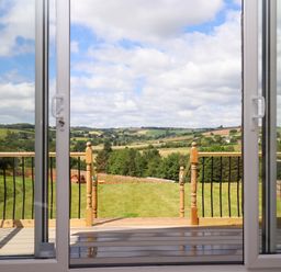 A view from sliding glass doors onto a wooden deck and a grassy yard with hills and fields in the distance at Ambleside in Cullompton
