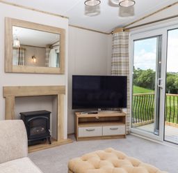 A living room with a large TV on a wooden stand a mirror above a fireplace and sliding glass doors leading to a balcony with a view of green fields at Ambleside in Cullompton