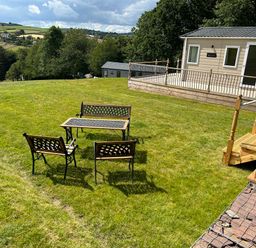 A grassy garden area with outdoor table and chairs near a raised deck and building at Ambleside in Cullompton
