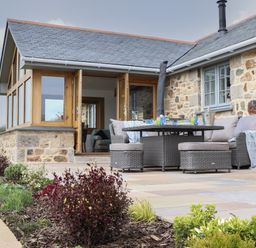 An outdoor patio with a round table and cushioned chairs next to a stone house with wooden framed glass doors and windows at Trevilges Cottage Trewennack near Helston