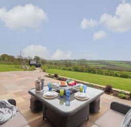 An outdoor patio area with a wicker dining table set with plates and glasses a grassy garden and countryside view at Trevilges Cottage in Trewennack near Helston