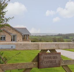 A wooden gate with a sign reading Trevilges Cottage with a stone and wood house and green fields in the background at Trevilges Cottage Trewennack near Helston