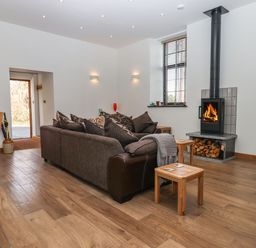 A living room with a brown sofa, wooden stools, a wood-burning stove, a dining table, and a window at The Old Chapel in Ilsington