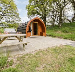 A small wooden pod shelter on a paved area with a picnic table and chairs surrounded by grass and trees at Conwy Pod in Capel Garmon near Betws-Y-Coed