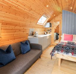 A wooden pod interior with a gray sofa and blue pillows a bed with pink pillows and folded towels a kitchenette with appliances and a skylight window at Conwy Pod in Capel Garmon near Betws-Y-Coed