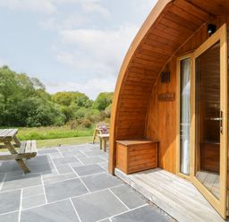 A wooden curved cabin with an open door on a stone patio with a wooden picnic table and benches outside Llugwy Pod Capel Garmon near Betws-Y-Coed