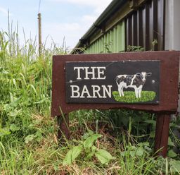A wooden sign with a painting of a cow and the words the barn in tall grass beside a metal building at Northwood Barn in Okehampton