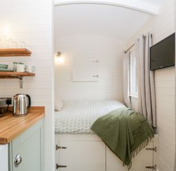 A small room with a bed, a wall-mounted TV, curtains, wooden shelves with dishes, a toaster, and a kettle at Willow in Kemble