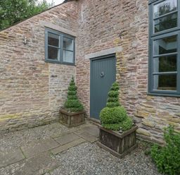 A stone building exterior with two windows a green door and two spiral-shaped topiary plants in wooden planters at Owl Barn Marston Stannett near Leominster