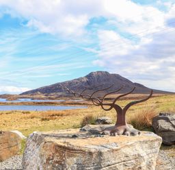 A metal sculpture resembling tree branches on a large rock with a mountain and lake in the background at North Light Studio Locheport near Lochmaddy North Uist