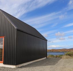 A black house with a red window frame beside a white building near a body of water at North Light Studio in Locheport near Lochmaddy North Uist