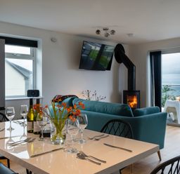 A dining table set with wine bottles and glasses next to a living room with a green sofa and a wood stove at Lanyon in St Ives