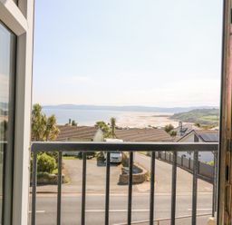 A view of a beach and houses from a window with a black balcony railing at Sea La Vie in Benllech