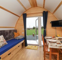 An interior view of a wooden pod with a blue cushioned bench, pillows, a dining table set for four, and a wall-mounted television at Mischief Little Kelk near Bridlington