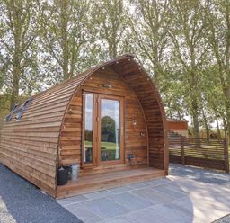 A wooden pod with a glass door and small porch on a paved area surrounded by trees at Mischief in Little Kelk near Bridlington