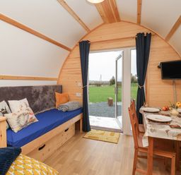 An interior view of a wooden pod with a blue cushioned bench, dining table with chairs, open door leading outside and a wall-mounted TV at Covey in Little Kelk near Bridlington