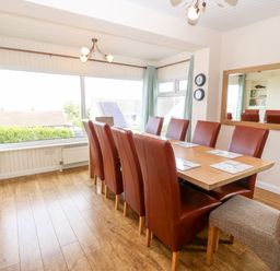 A dining room with a wooden table surrounded by red and beige chairs next to large windows at Willoway in Benllech