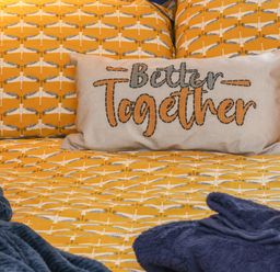 A bed with yellow patterned bedding a cushion with better together text and blue towels