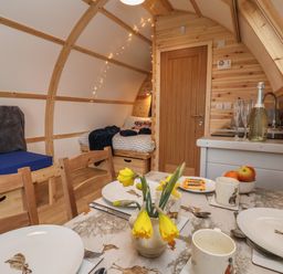 An interior of a small wooden cabin with a dining table set with plates and cups a small kitchenette a door a bed with blankets and pillows and a bench seat at Parliament in Little Kelk near Bridlington
