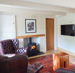 A living room with two leather armchairs a wood stove a wooden side table a wall-mounted television and a red rug at Spring Garden Cottage in Laugharne