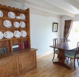 A dining room with a wooden table and chairs and a wooden cabinet with plates and books at Spring Garden Cottage in Laugharne