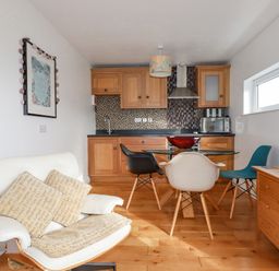 A kitchen with wood cabinets a glass dining table with three chairs a white armchair with cushions and a wooden side table with lamp at Lapwing in Perranporth