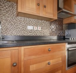 A kitchen countertop with a sink, wooden cabinets, a built-in oven, a range hood, a microwave, and a tiled backsplash at Lapwing in Perranporth
