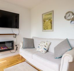 A sitting room with a wall mounted TV a wall clock a floor lamp and a sofa with cushions at Lapwing in Perranporth