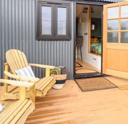 An outdoor wooden deck with two wooden chairs with cushions a basket with blankets an open wooden door leading to an interior space at Portland Luxury Glamping in Nottingham