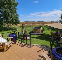 A wooden deck with Adirondack chairs and blue pots overlooking a grassy yard with a hot tub a shaded seating area and barbecue at Portland Luxury Glamping in Nottingham