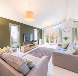 A living room with two sofas a large rug a wall mounted TV and glass doors at Fern Lodge in Allithwaite near Cartmel