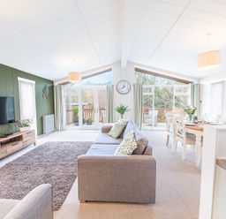 A living room with a grey sofa and a dining table with chairs near large windows with green curtains at Fern Lodge in Allithwaite near Cartmel