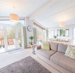 A living room with a sofa dining table plants and large windows at Fern Lodge in Allithwaite near Cartmel